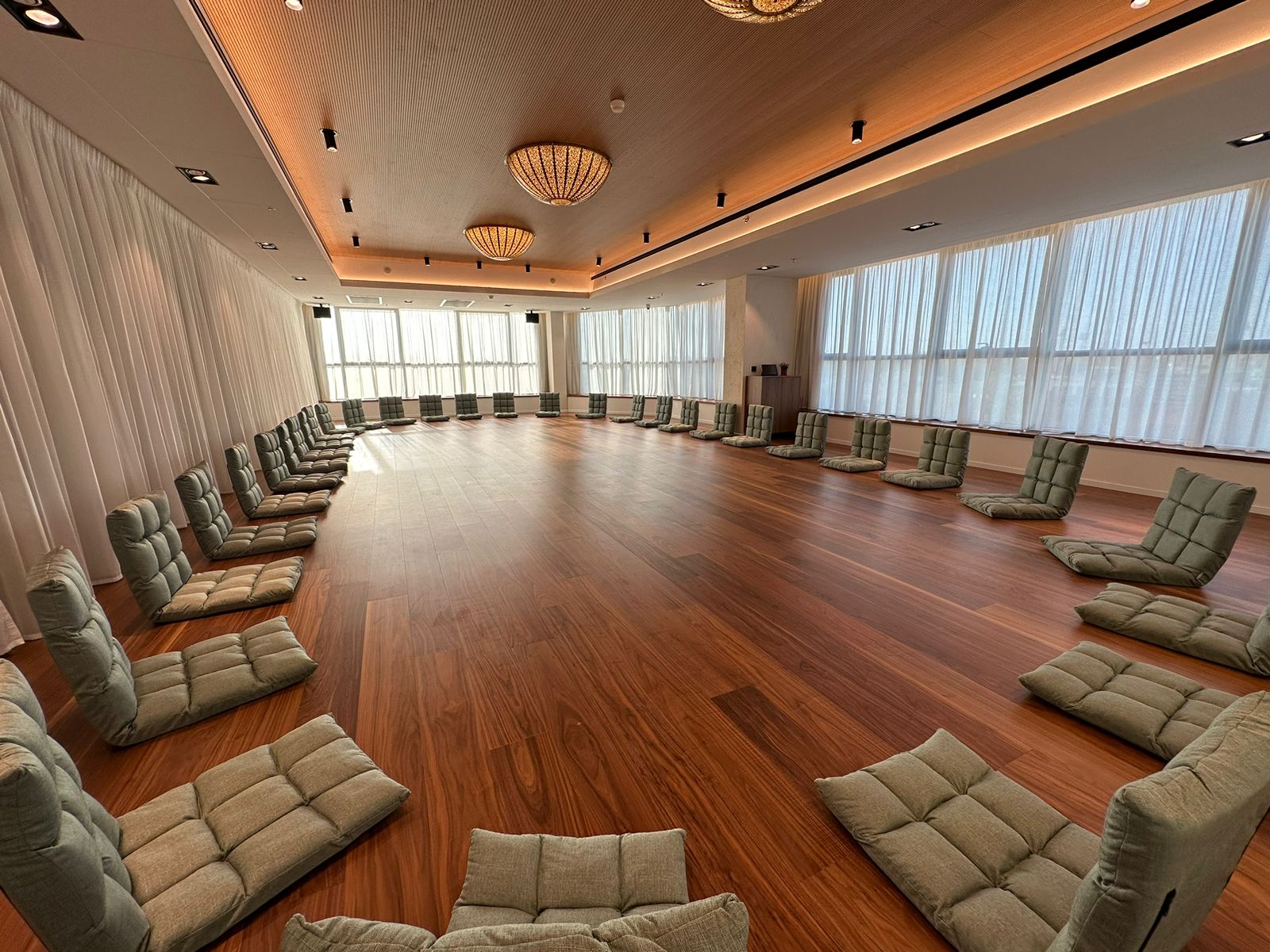 Spacious room with wooden floor and a large circular seating arrangement of gray cushioned floor chairs under warm ceiling lights.