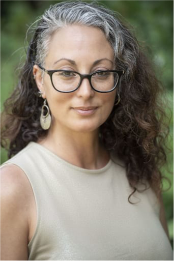 אישה עם שיער מתולתל אפור ושחור, משקפיים גדולות ועגילים עגולים, לובשת חולצת שרוולים קצרה בהירה עם רקע ירוק מטושטש.