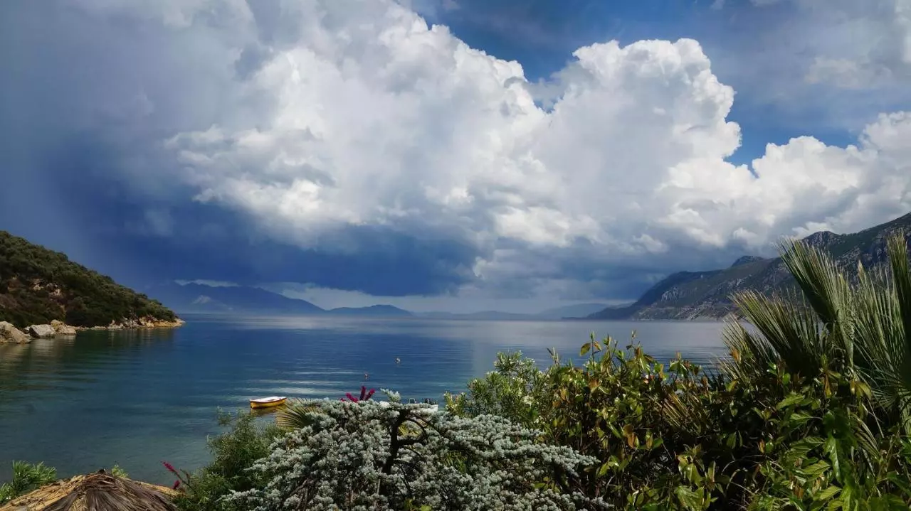 שמיים עם עננים גדולים מעל אגם או ים שקוע מוקף בצמחייה ירוקה והרים ברקע.