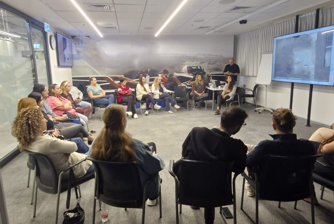 קבוצה של אנשים יושבים בחדר מעגלי בסדרות, צופים במסך גדול בזמן הרצאה או דיון.