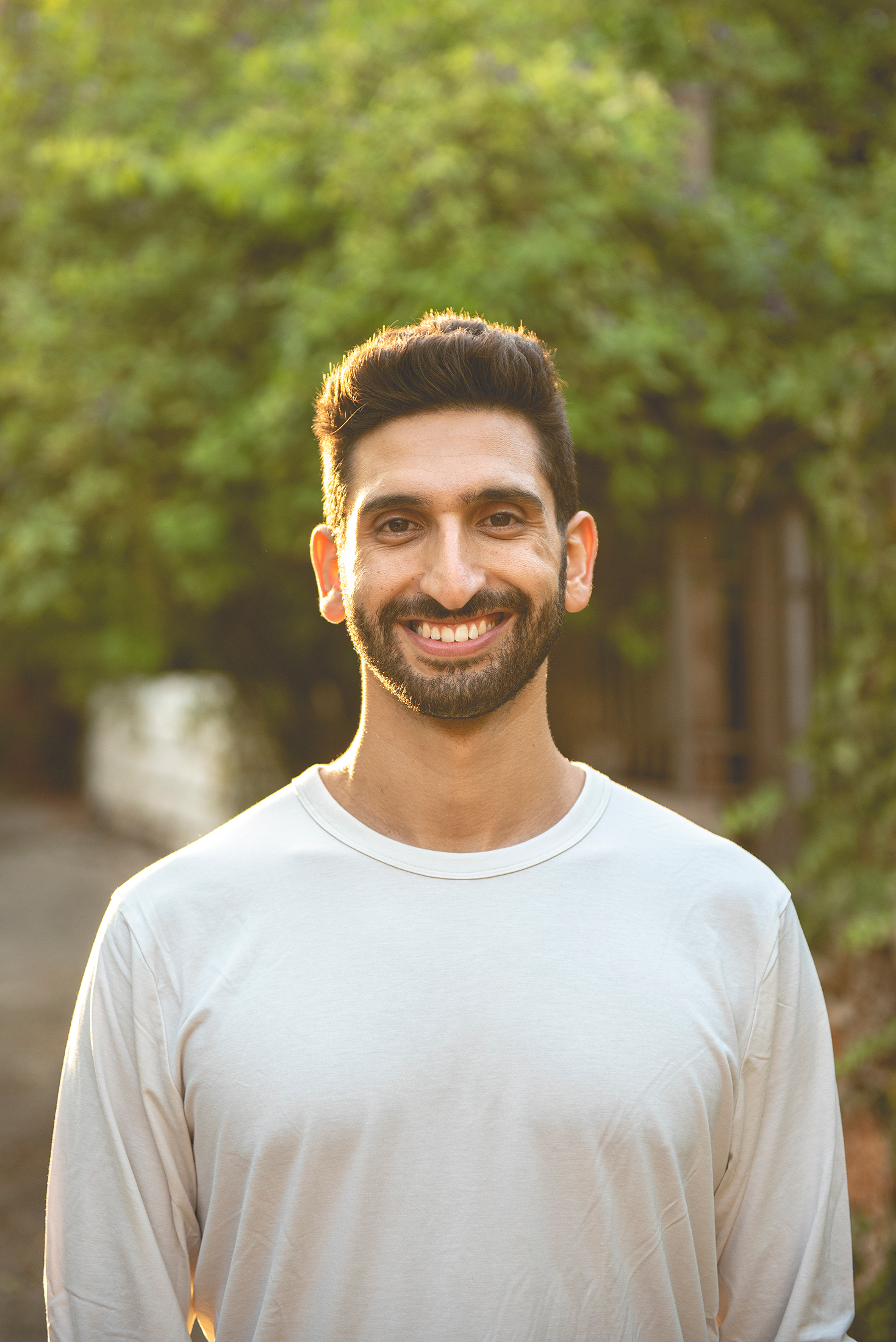 גבר צעיר מחייך עם זקן קצר וחולצה לבנה, ברקע עצים ושטח פתוח מטושטש.