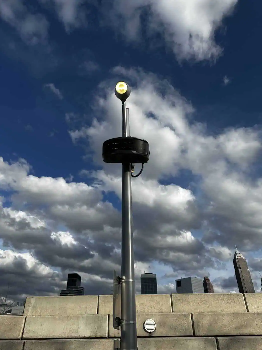 ROSA Edge AI security camera mounted on a light pole against a cloudy sky and city skyline