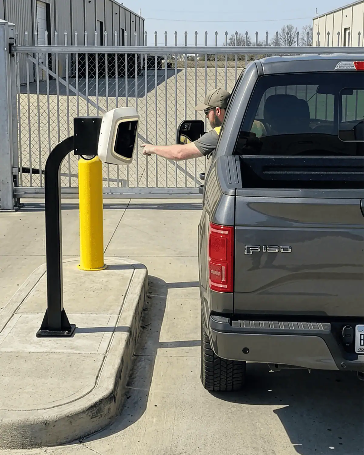 Driver in a pickup reaching to an AVA Gen4 unit on a gooseneck pedestal at a warehouse gate, seen from behind the truck