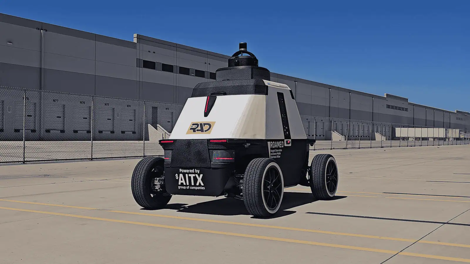 ROAMEO patrol robot in a distribution center truck yard under a clear blue sky, seen from behind