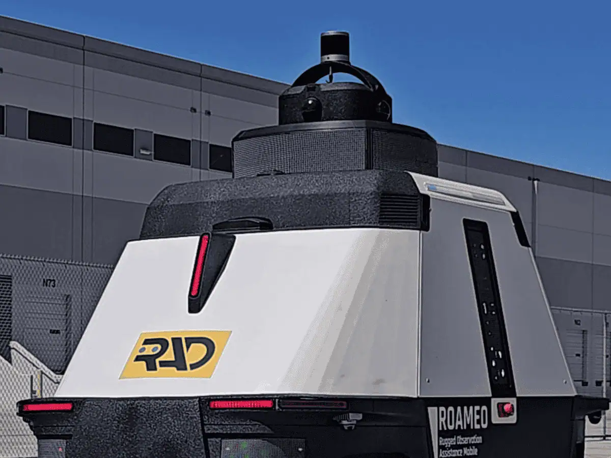 Sensor mast on top of a ROAMEO patrol robot, with a LiDAR puck above a ring of cameras, outside a distribution center
