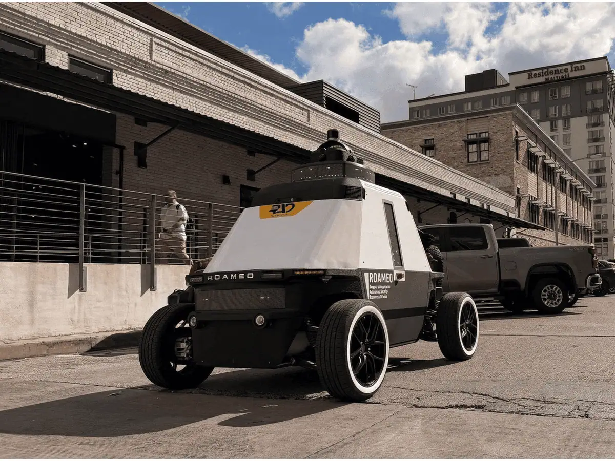 ROAMEO patrol robot on a city street beside a loading dock, a parked pickup, and a pedestrian