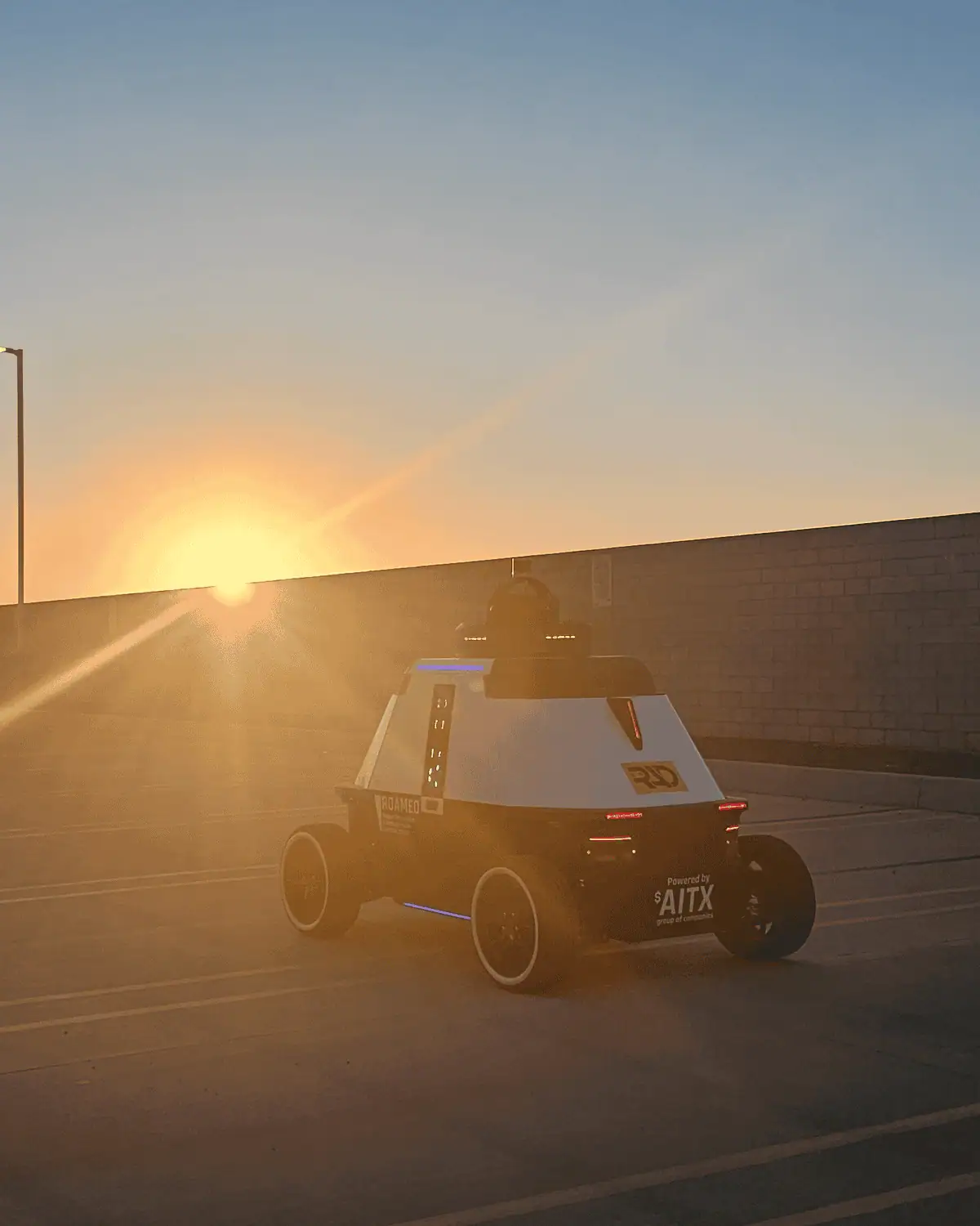 ROAMEO patrol robot on a rooftop parking deck at sunset, seen from behind with the sun low over a wall
