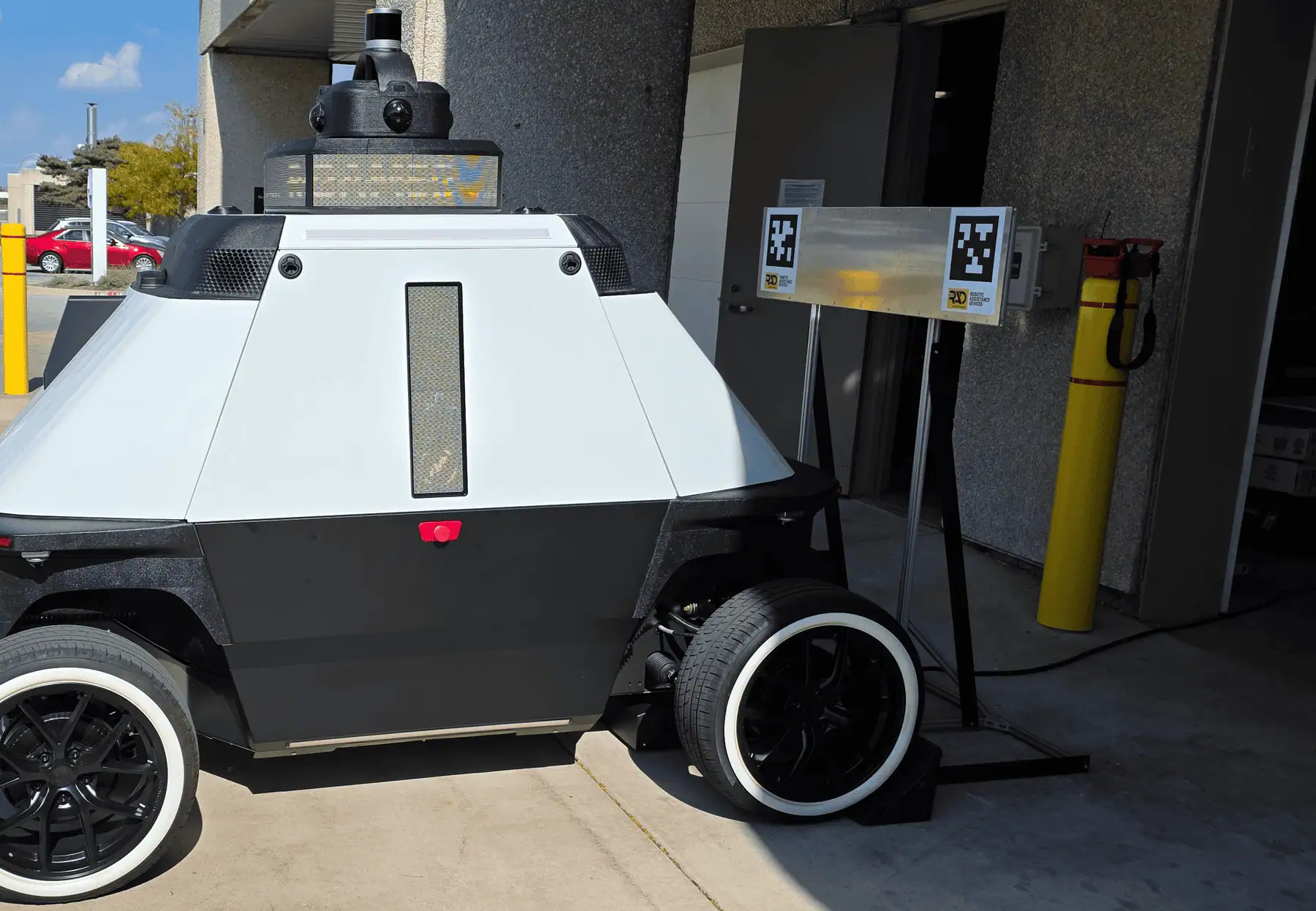 ROAMEO parked beside its charging dock and fiducial marker board at a building entrance, seen from behind in daylight