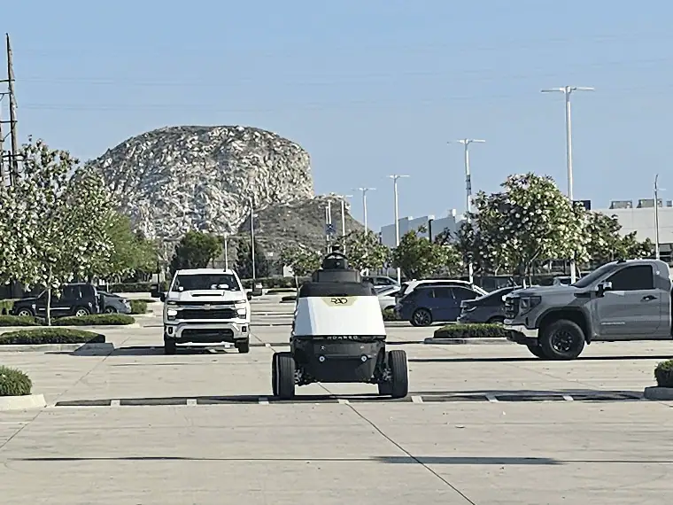 ROAMEO patrolling down an open lane of a daytime parking lot between parked pickups, with a rocky hill behind