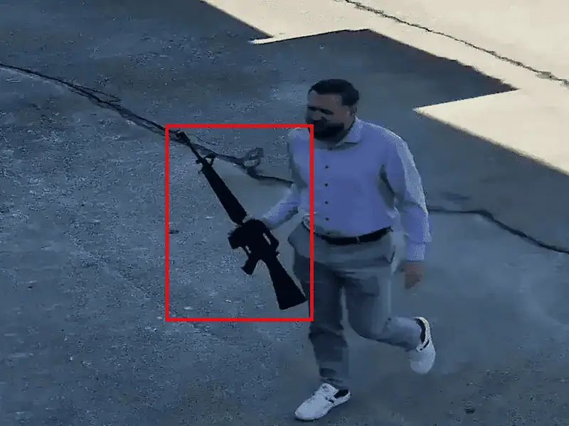 Staged test frame of a man carrying a rifle across a parking lot, the rifle outlined in a red detection box
