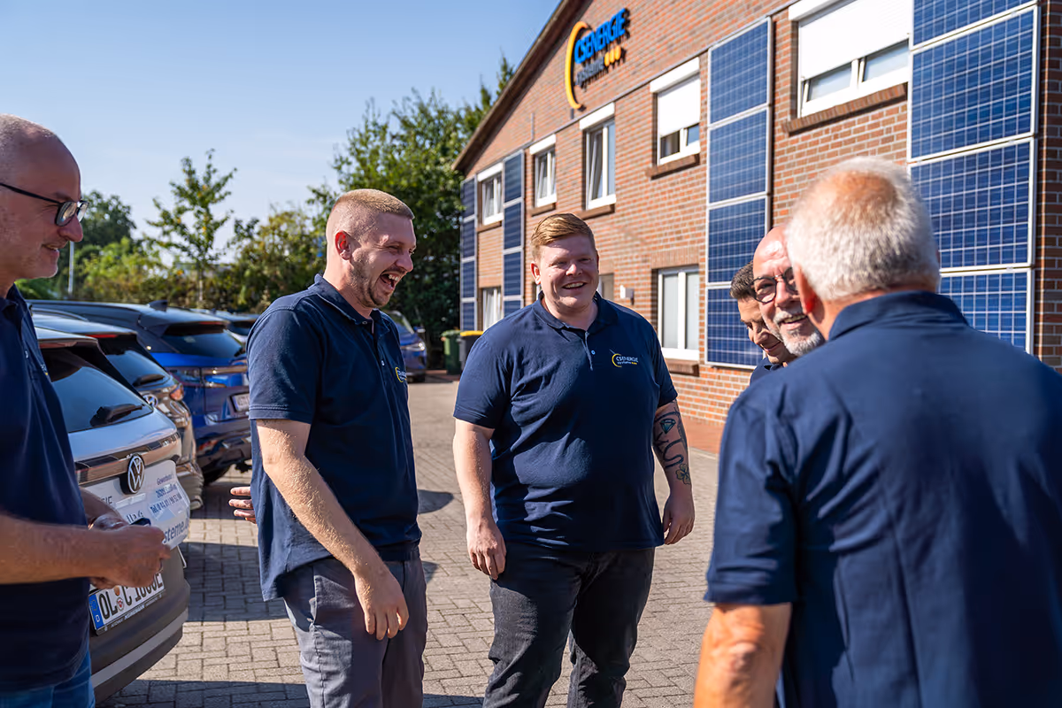 Fünf Männer in blauen Poloshirts mit Firmenlogo unterhalten sich und lachen vor einem Gebäude mit Solarpaneelen an der Fassade.