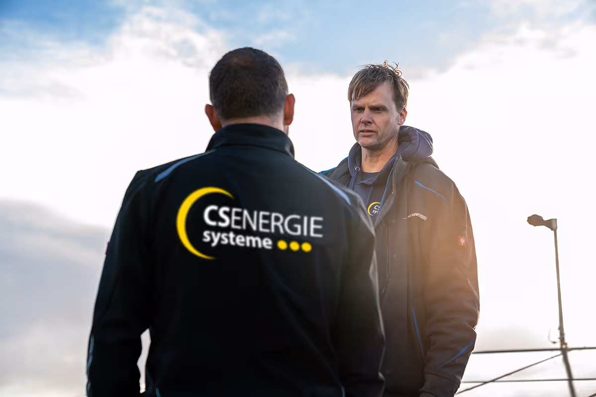 Zwei Männer in Arbeitskleidung mit CS Energie Systeme-Logo im Freien unter bewölktem Himmel.