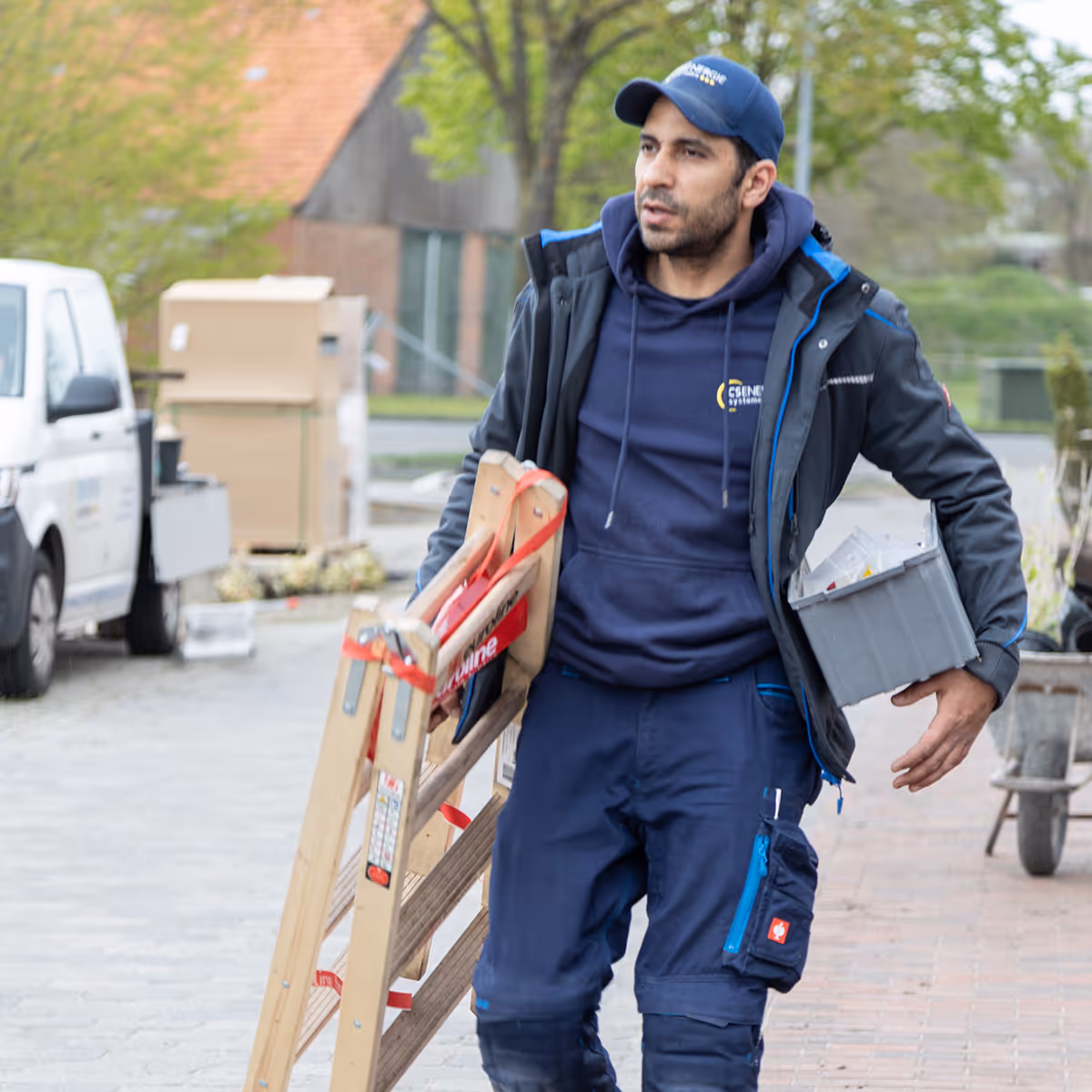 Handwerker trägt eine Leiter in der einen und eine graue Kiste in der anderen Hand auf einem Bürgersteig.