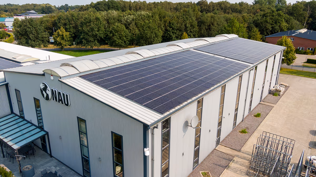 Moderne Industriehalle mit Solarzellen auf dem Dach und einem NAU-Logo an der Fassade, umgeben von Bäumen.