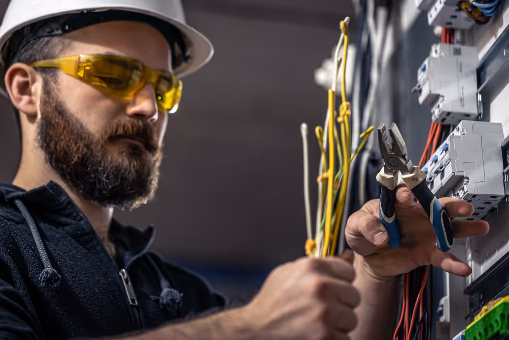 Elektriker mit Helm und Schutzbrille arbeitet an einem Schaltschrank mit Zangen und mehreren gelben Kabeln.