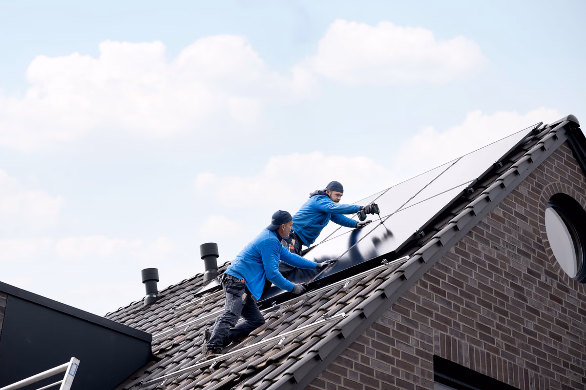 Zwei Arbeiter in blauen Jacken installieren Solarmodule auf einem geneigten Ziegeldach eines Hauses unter bewölktem Himmel.