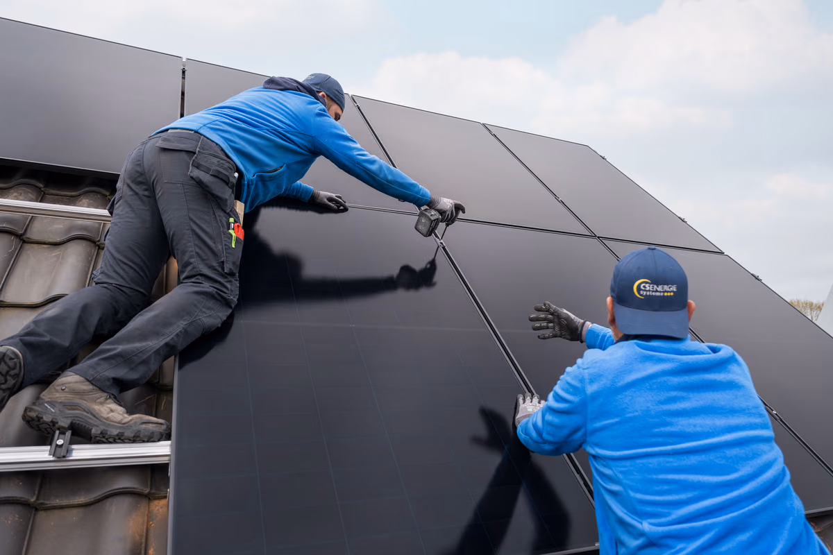Zwei Arbeiter montieren schwarze Solarmodule auf einem Dach bei bewölktem Himmel.