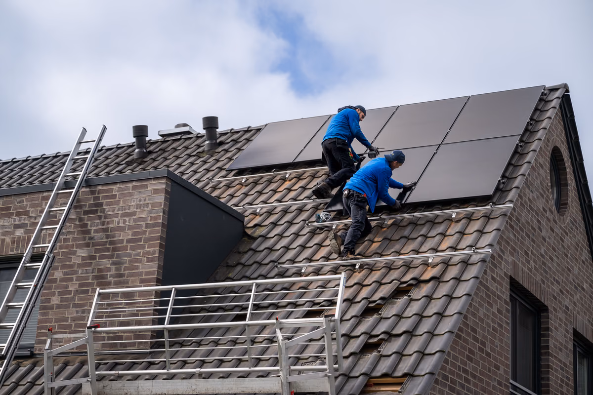 Zwei Handwerker in blauen Jacken installieren Solarpaneele auf dem Dach eines Hauses mit dunklen Dachziegeln unter einem bewölkten Himmel.