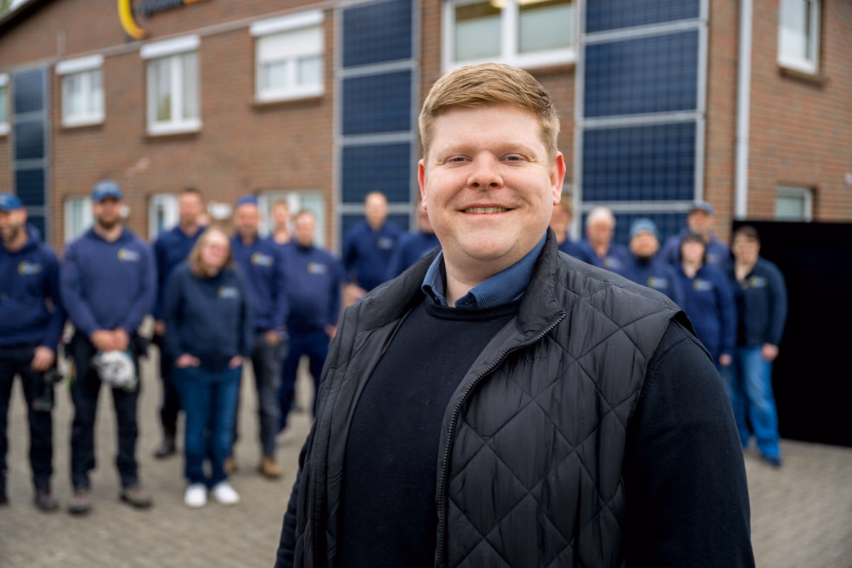 Ein lächelnder Mann mit schwarzer gesteppter Weste und blauem Hemd steht im Vordergrund, im Hintergrund steht eine Gruppe von Menschen in blauen Arbeitskleidern vor einem Gebäude mit Solarpaneelen an der Fassade.