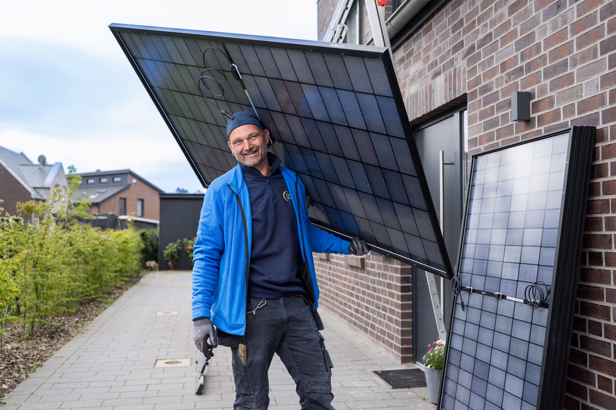 Ein lächelnder Mann in blauer Jacke und schwarzer Mütze trägt ein großes Solarpanel neben einem Backsteingebäude auf einem Gehweg mit weiteren Häusern und Pflanzen im Hintergrund.