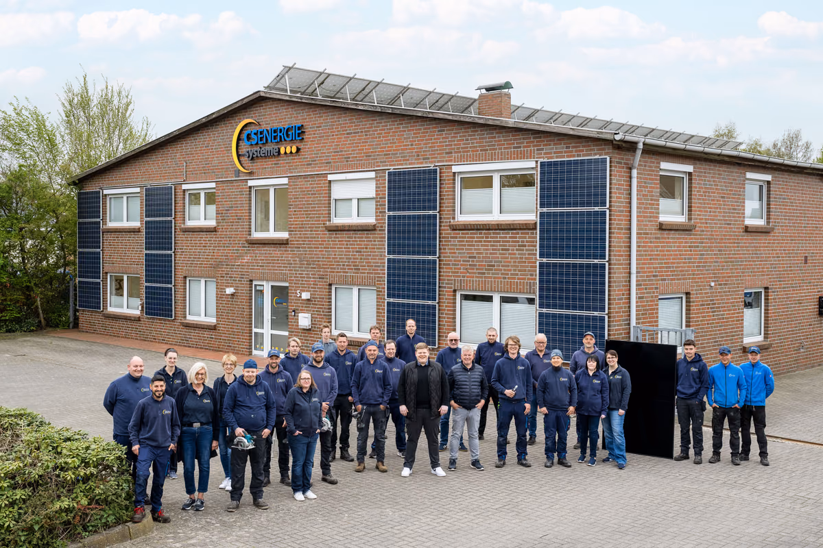 Gruppenfoto von 24 Personen vor einem Backsteingebäude mit Solarmodulen an der Fassade und einem Schild mit der Aufschrift GS ENERGIE systeme, die meisten tragen blaue Arbeitskleidung.