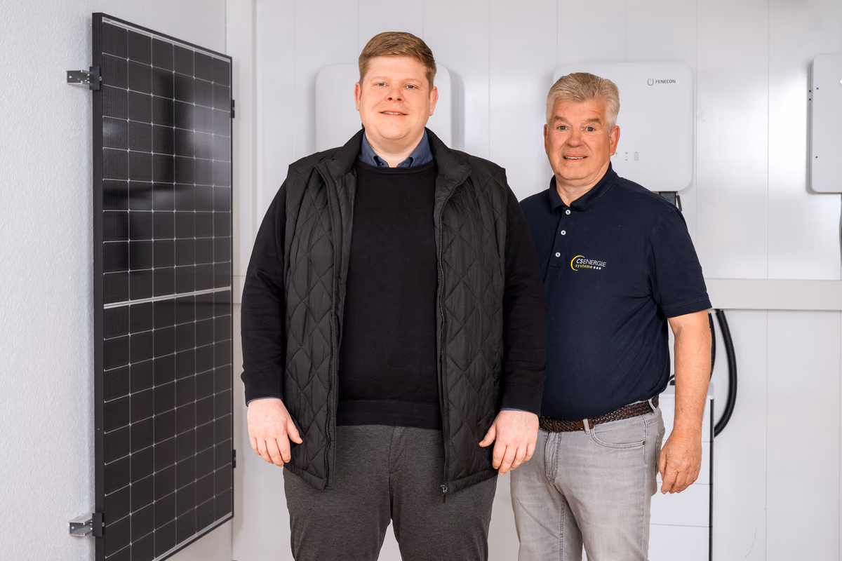 Zwei Männer stehen nebeneinander in einem Raum mit weißen Wänden, im Hintergrund sind Solarpaneele und technische Geräte an der Wand montiert.