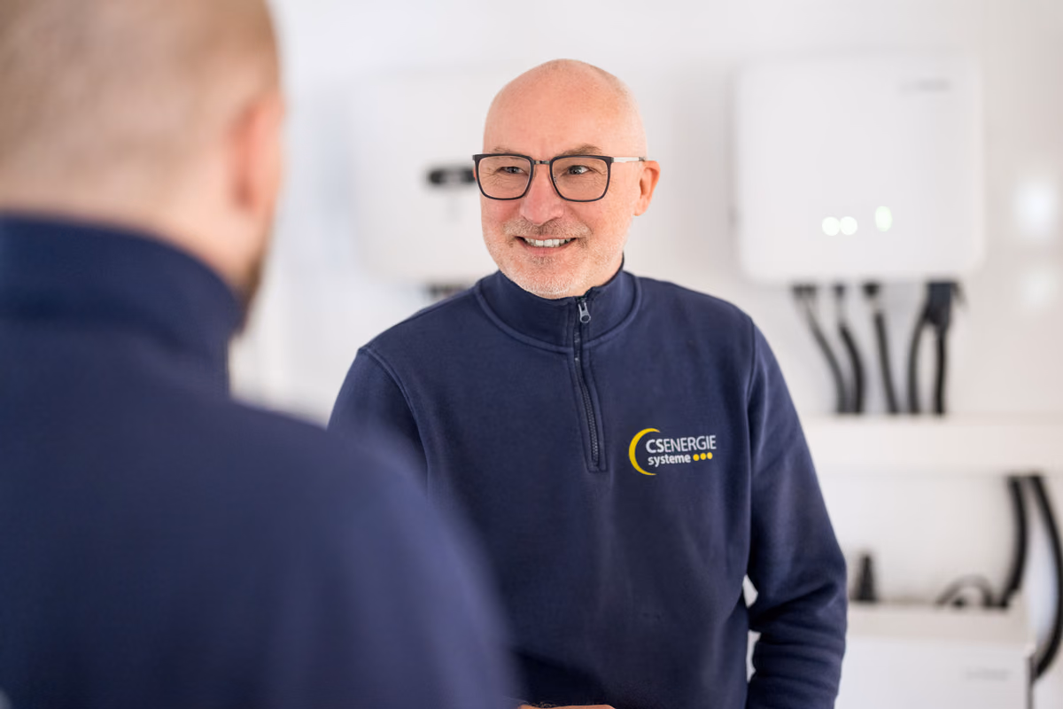Älterer Mann mit Brille und blauem Pullover mit Csenergie Systeme-Logo, der lächelnd in einem Raum mit elektrischen Geräten und Kabeln steht.