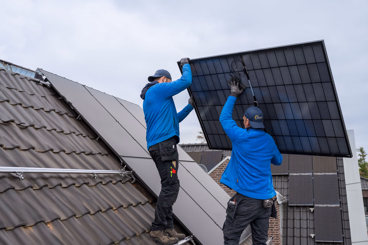 Zwei Arbeiter in blauen Jacken und Handschuhen installieren ein großes Solarpanel auf einem geneigten Dach mit braunen Ziegeln unter bewölktem Himmel.