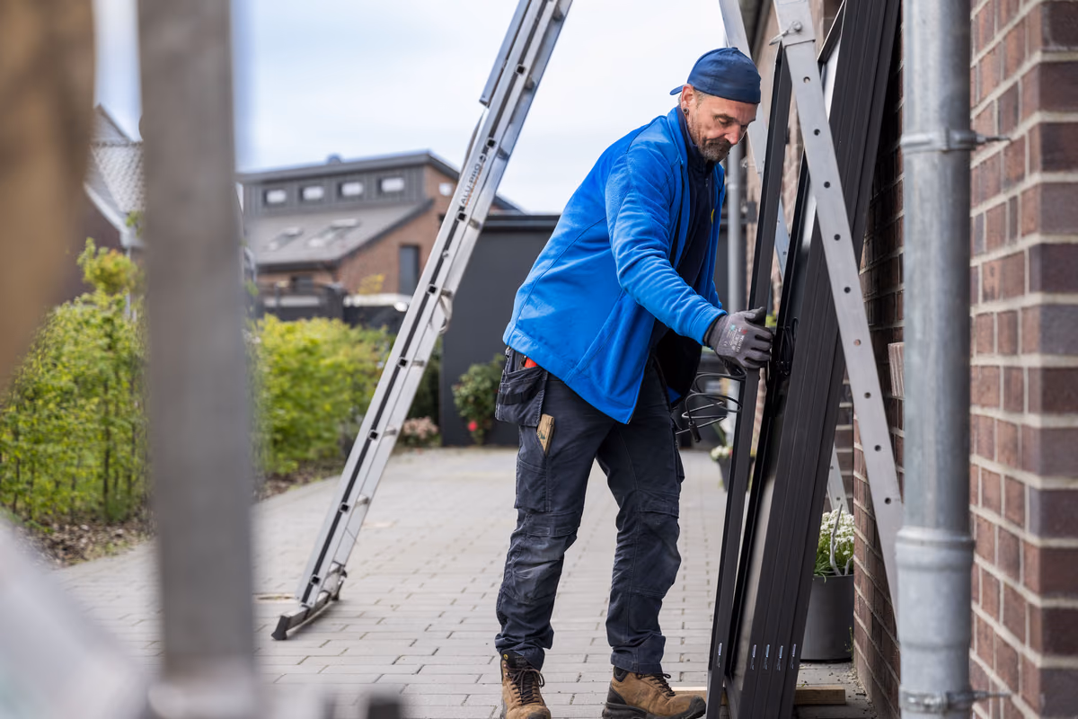 Ein Mann in blauer Jacke und schwarzer Mütze montiert schwarze Paneele an der Backsteinwand eines Hauses, während eine Leiter neben ihm steht.
