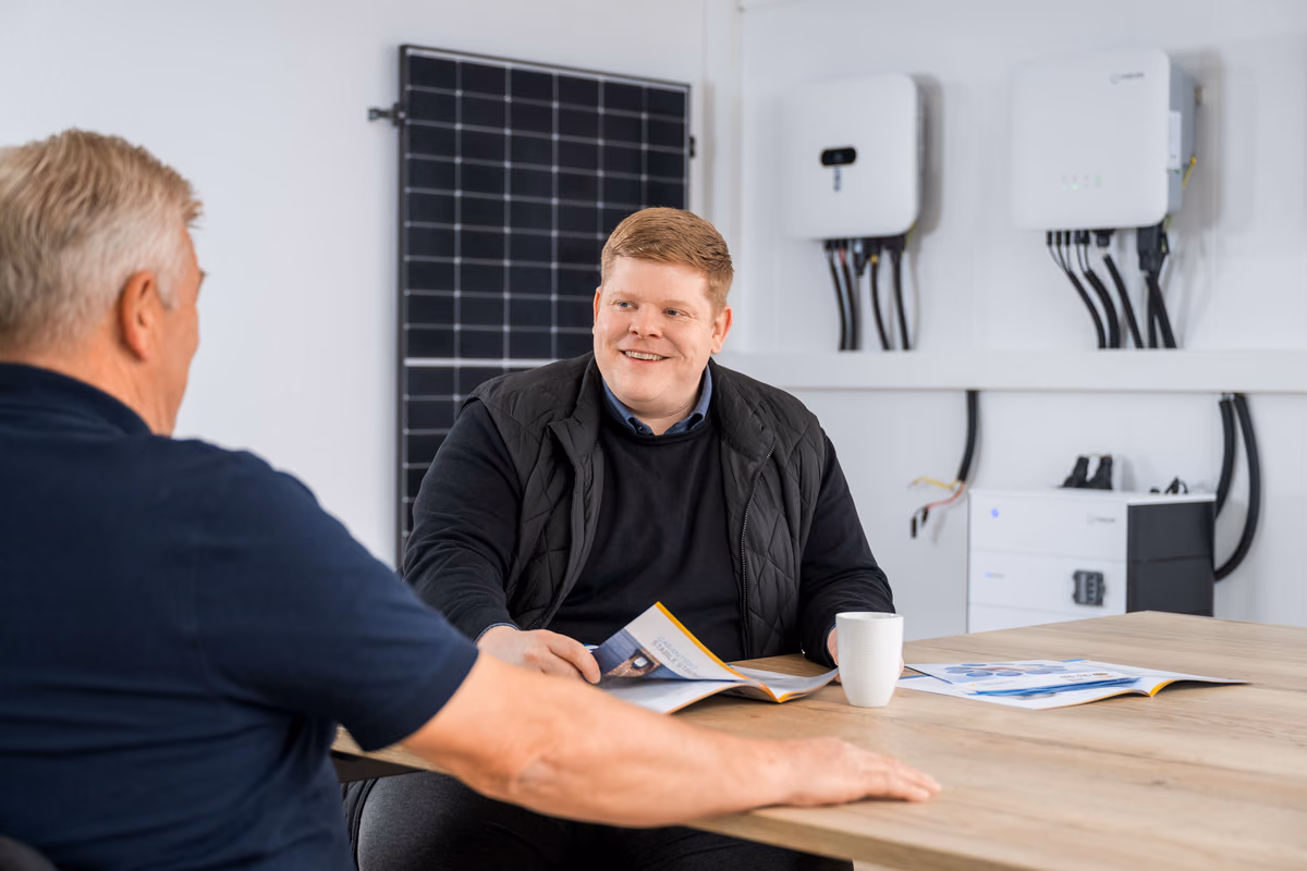 Zwei Männer sitzen an einem Holztisch in einem modernen Büro mit Solarmodulen und technischen Geräten an der Wand; einer liest eine Broschüre und lächelt.