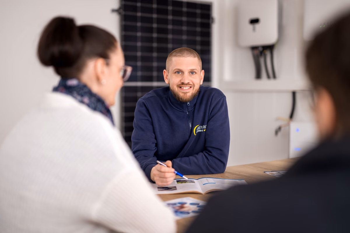 Ein Mann in einer marineblauen Jacke mit dem Logo 'GENROCK' sitzt an einem Tisch und lächelt, während er mit zwei Personen spricht, die im Vordergrund unscharf zu sehen sind. Im Hintergrund befindet sich eine Wand mit einem Solarpanel und elektrischen Geräten.