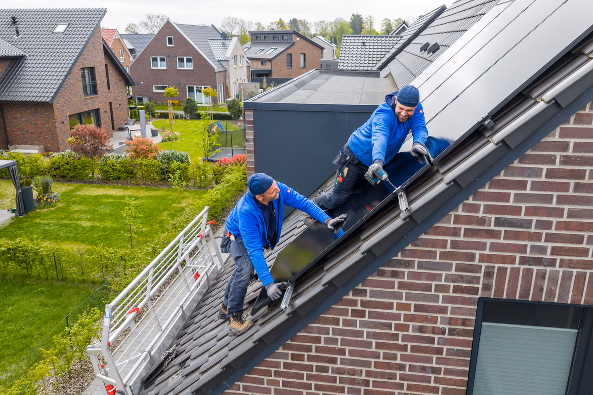 Zwei Handwerker in blauen Jacken montieren Solarpaneele auf einem geneigten Dach in einer Wohngegend mit modernen Häusern.