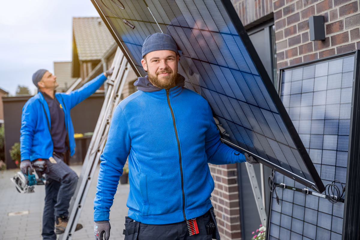 Zwei Arbeiter in blauen Jacken installieren Solarmodule an einer Hauswand aus Ziegeln, einer hält ein Solarpanel, der andere steht auf einer Leiter mit Werkzeug.