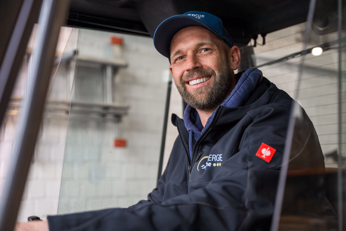 Lächelnder Mann mit Jacke, Kappe und Bart sitzt im Fahrzeug in einer Werkstatt oder Lagerhalle.