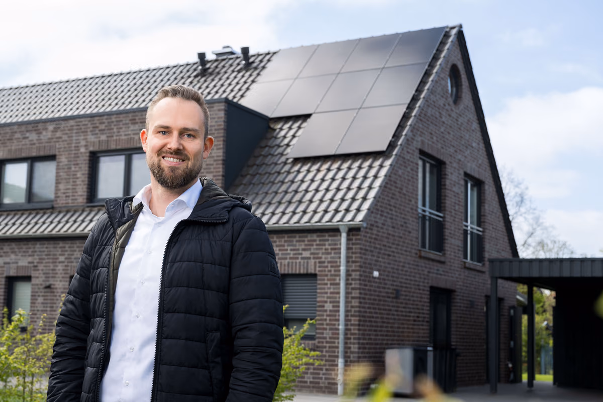 Mann mit Bart und schwarzer Jacke steht vor einem modernen Haus aus dunklem Ziegelstein mit Solarpaneelen auf dem Dach an einem bewölkten Tag.