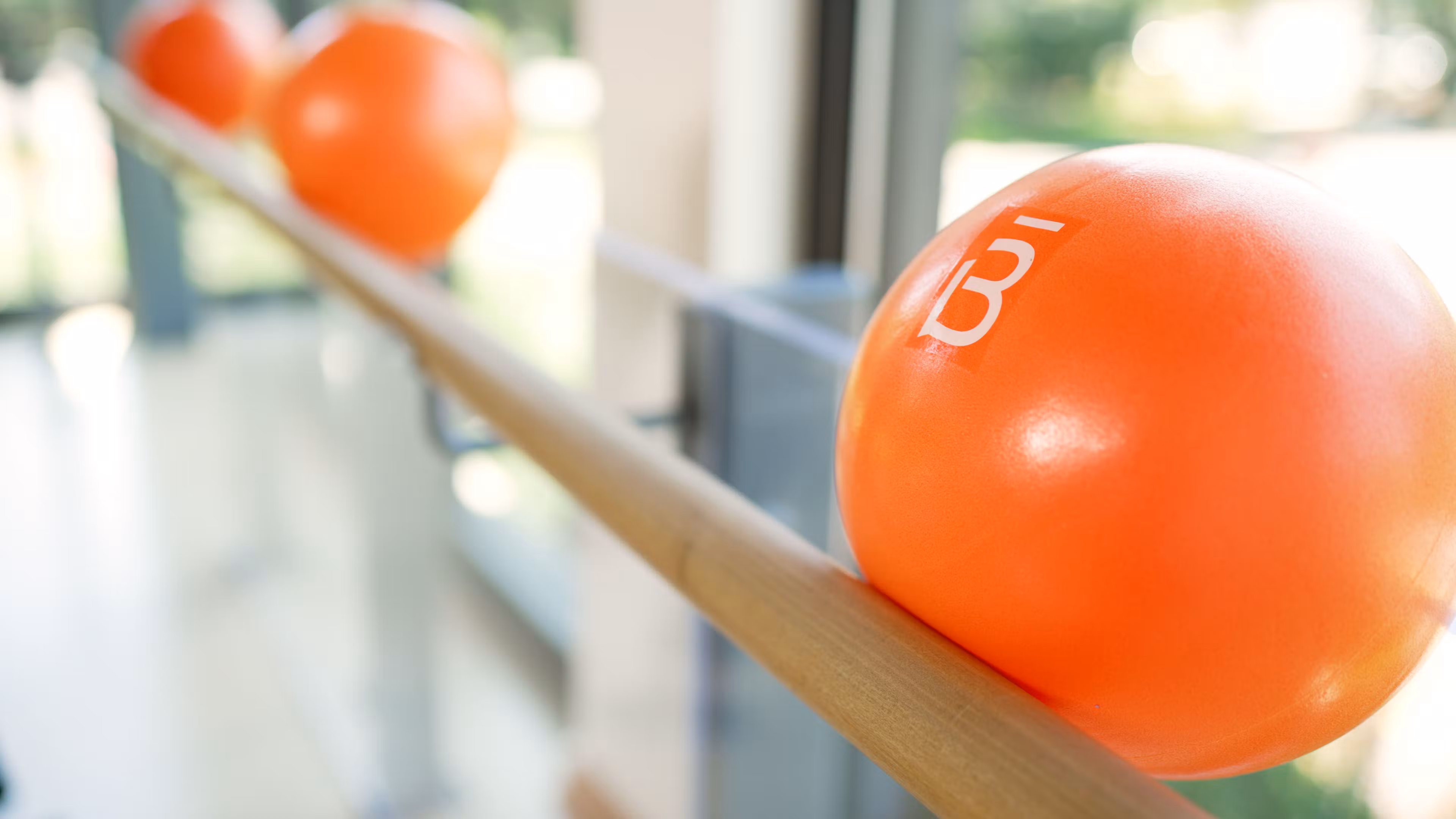Primer plano de una pelota de ejercicio barr3 apoyada sobre una barra de ballet de madera con un fondo borroso de otras dos pelotas naranjas en barre3 Santiago.
