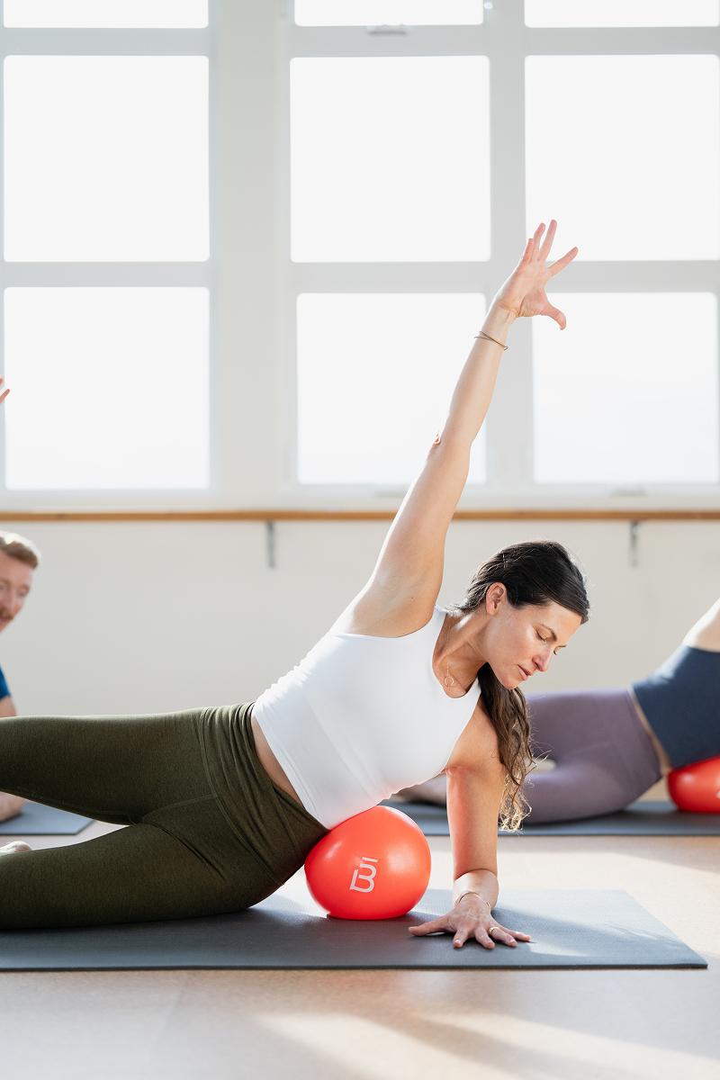 Mujer con camiseta blanca haciendo postura de plancha lateral de yoga sobre una colchoneta, usando una pelota de ejercicio barre3 debajo de su costado.