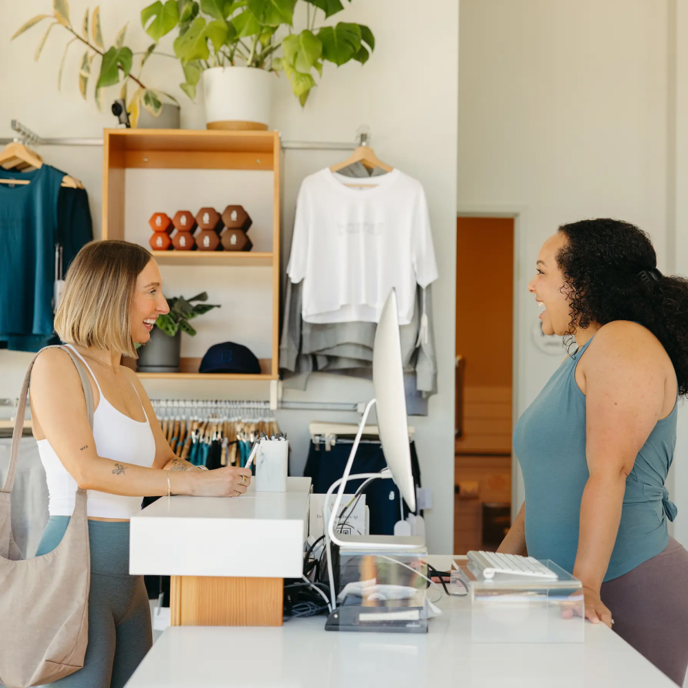 Dos mujeres sonriendo y hablando en el mostrador de una caja registradora, con ropa y mancuernas expuestas detrás de ellas en barre3 Santiago.