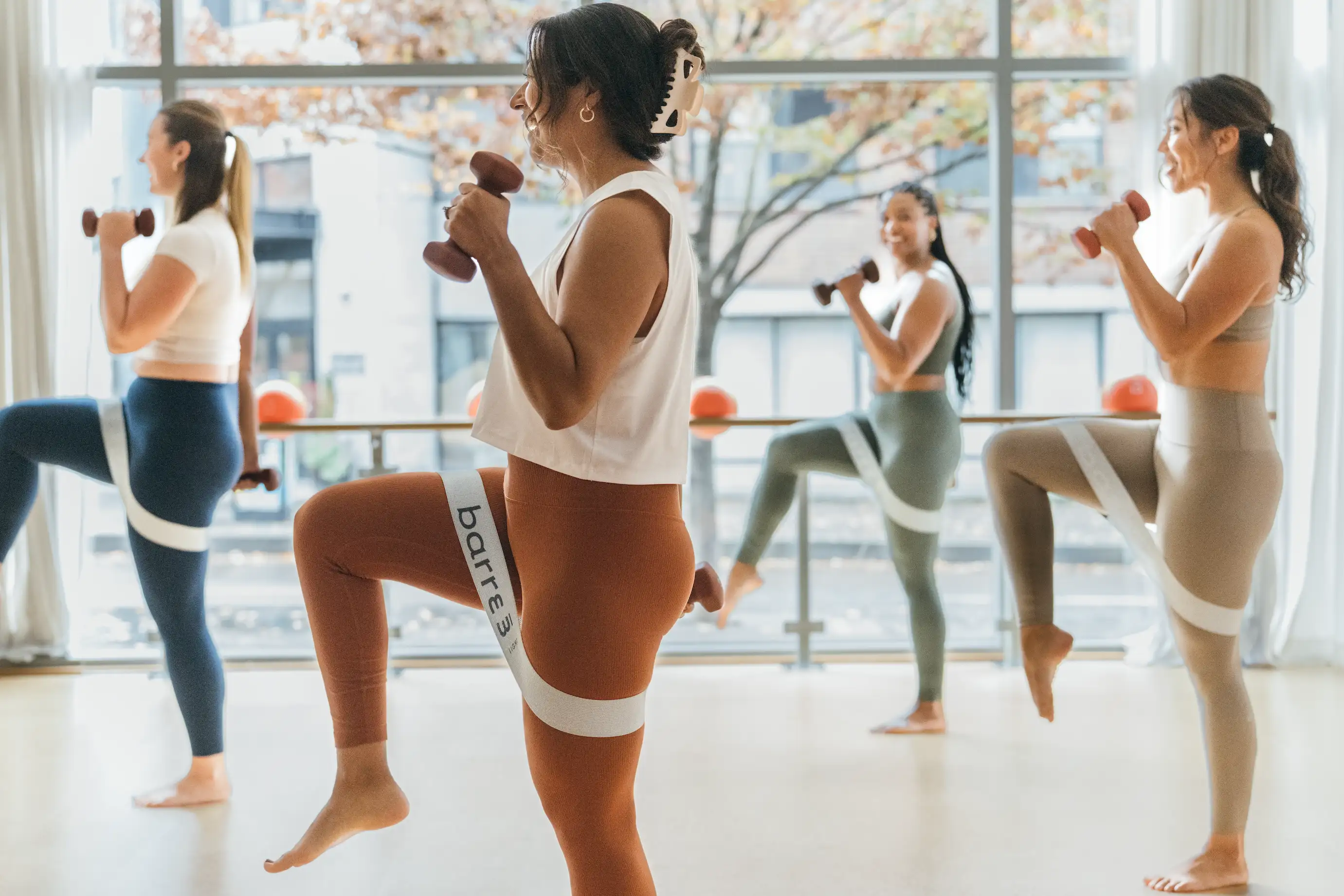 Cuatro mujeres se ejercitan en barre3 Santiago con bandas de resistencia alrededor de los muslos y sosteniendo pequeñas mancuernas, levantando las rodillas en una clase de fitness barre3.