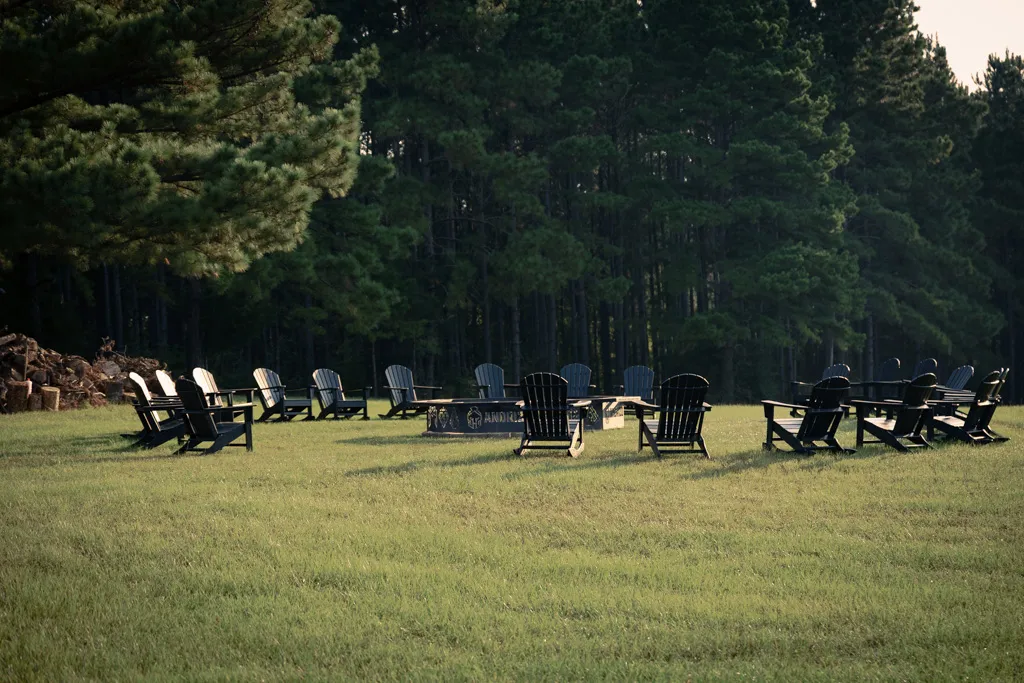 Circle of Adirondack chairs arranged around a fire pit on green grass with a dense pine forest in the background.