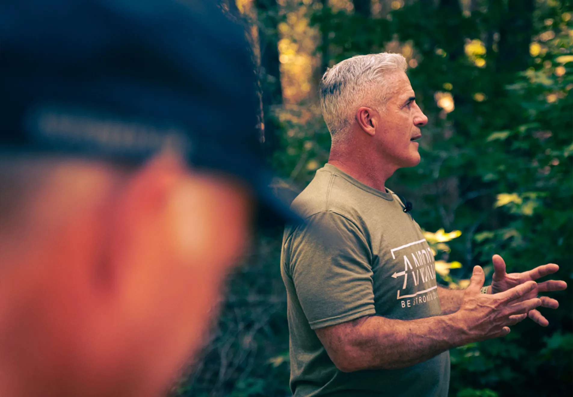 James deMelo in an olive green t-shirt speaking outdoors with blurred person in foreground.