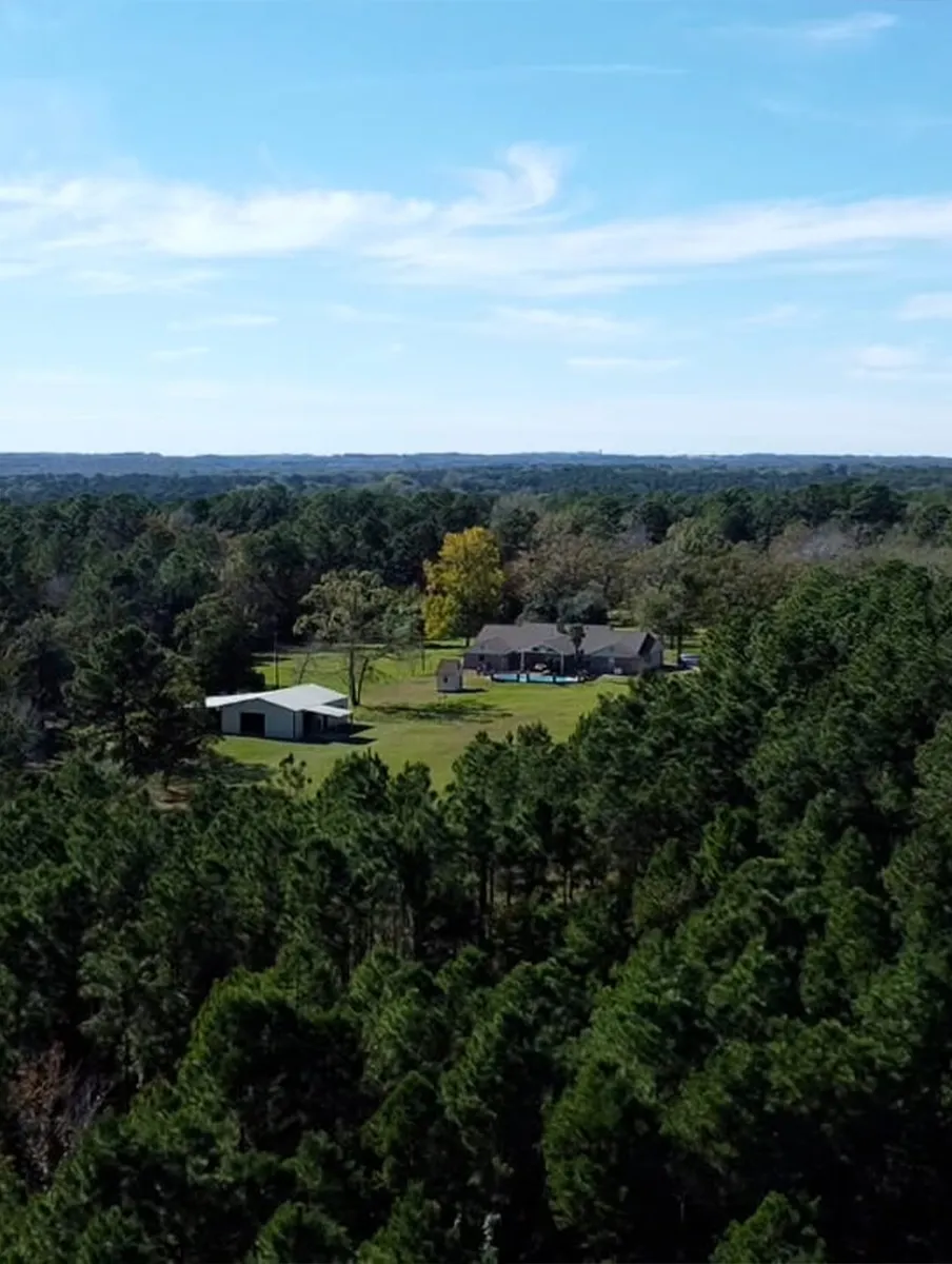 A rural ranch house on a large serene property, surrounded by dense pine forest under a clear blue sky.