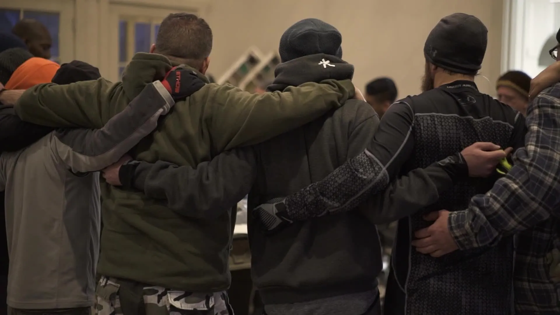 Group of men standing close with arms around each other's shoulders in a show of camaraderie indoors.