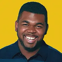 Smiling man with short hair and beard wearing a dark blue shirt against a yellow background.