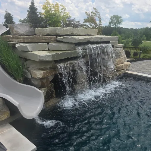 Stone waterfall feature flowing into a pool with a gray slide on the left side and trees in the background under a partly cloudy sky.