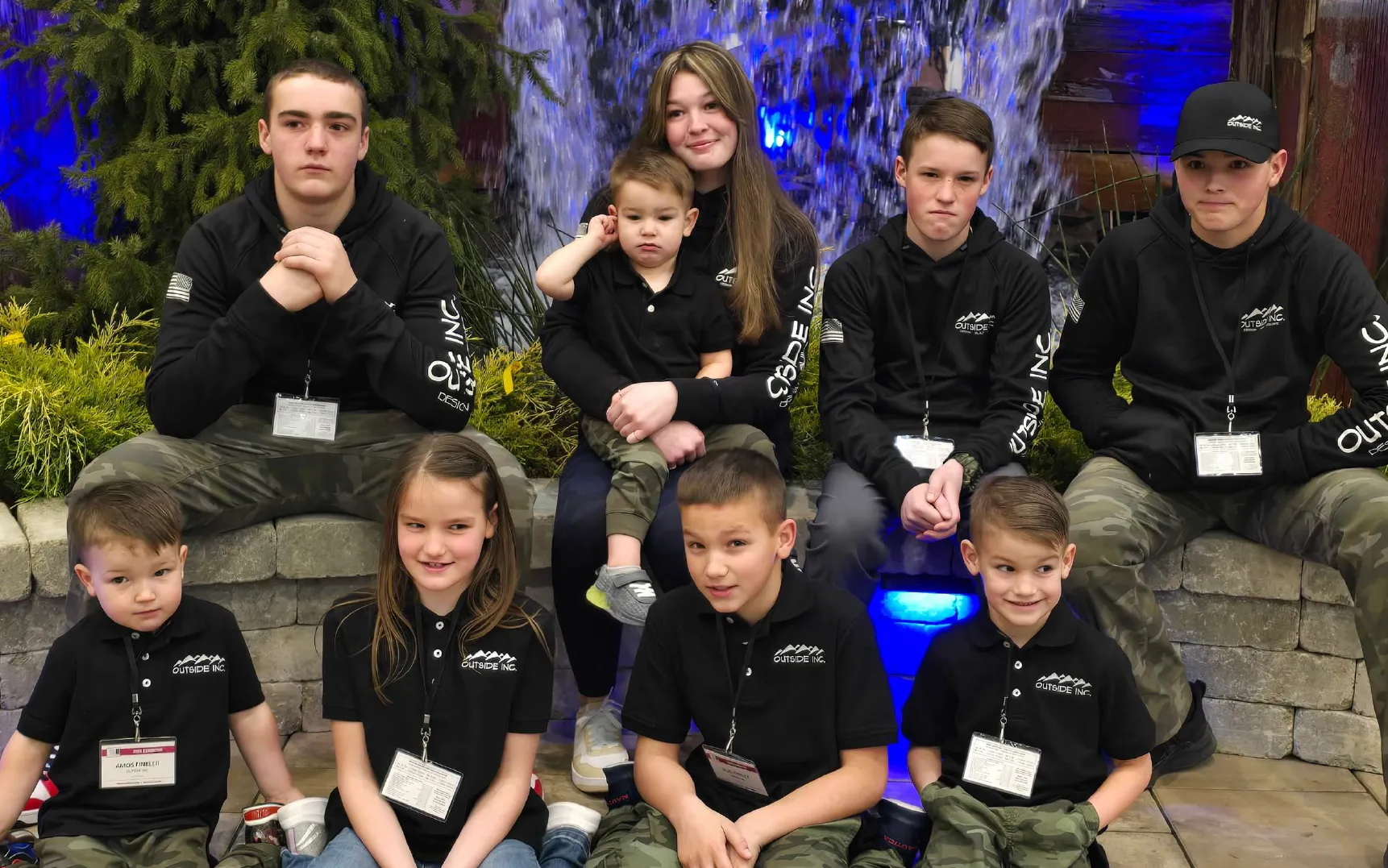 Group of children and teenagers wearing matching Outside Inc. shirts and badges, sitting and posing in front of a stone wall with greenery and a waterfall feature with blue lighting.