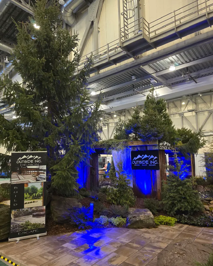 Indoor landscaping display with large pine trees, greenery, rocks, and blue lighting under a high industrial ceiling.