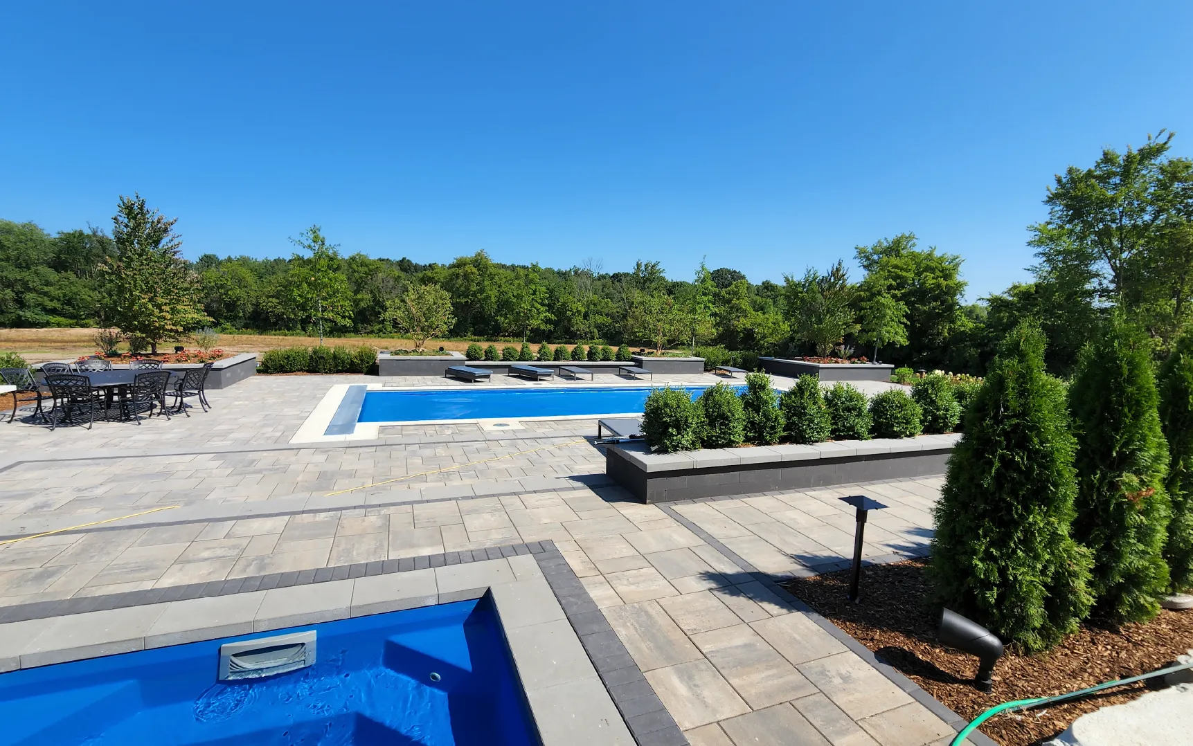 Modern outdoor patio with a swimming pool, hot tub, lounge chairs, dining table, and surrounding greenery under a clear blue sky.