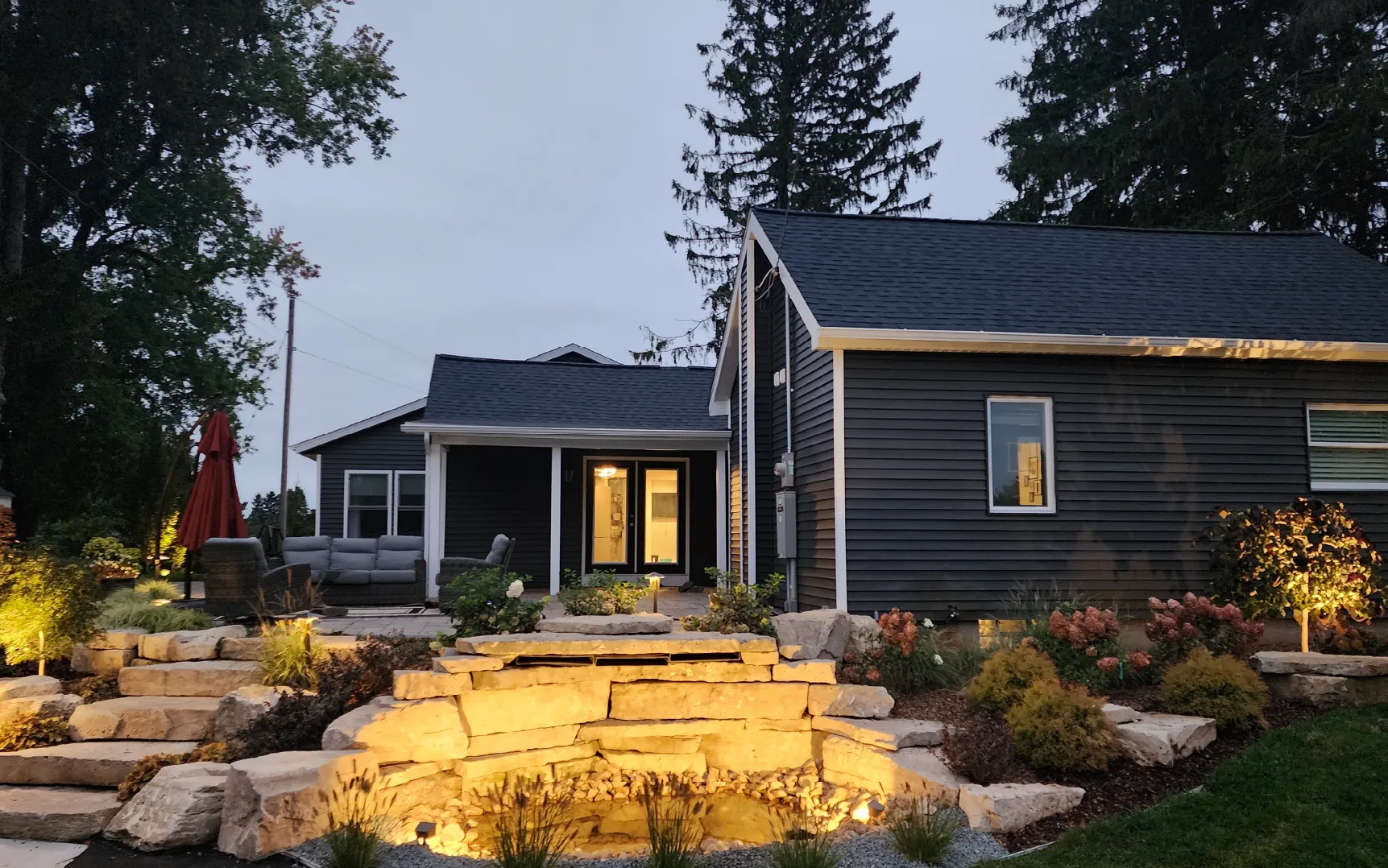 Evening view of a dark gray house with an illuminated stone water feature and a patio with outdoor seating and a red umbrella.