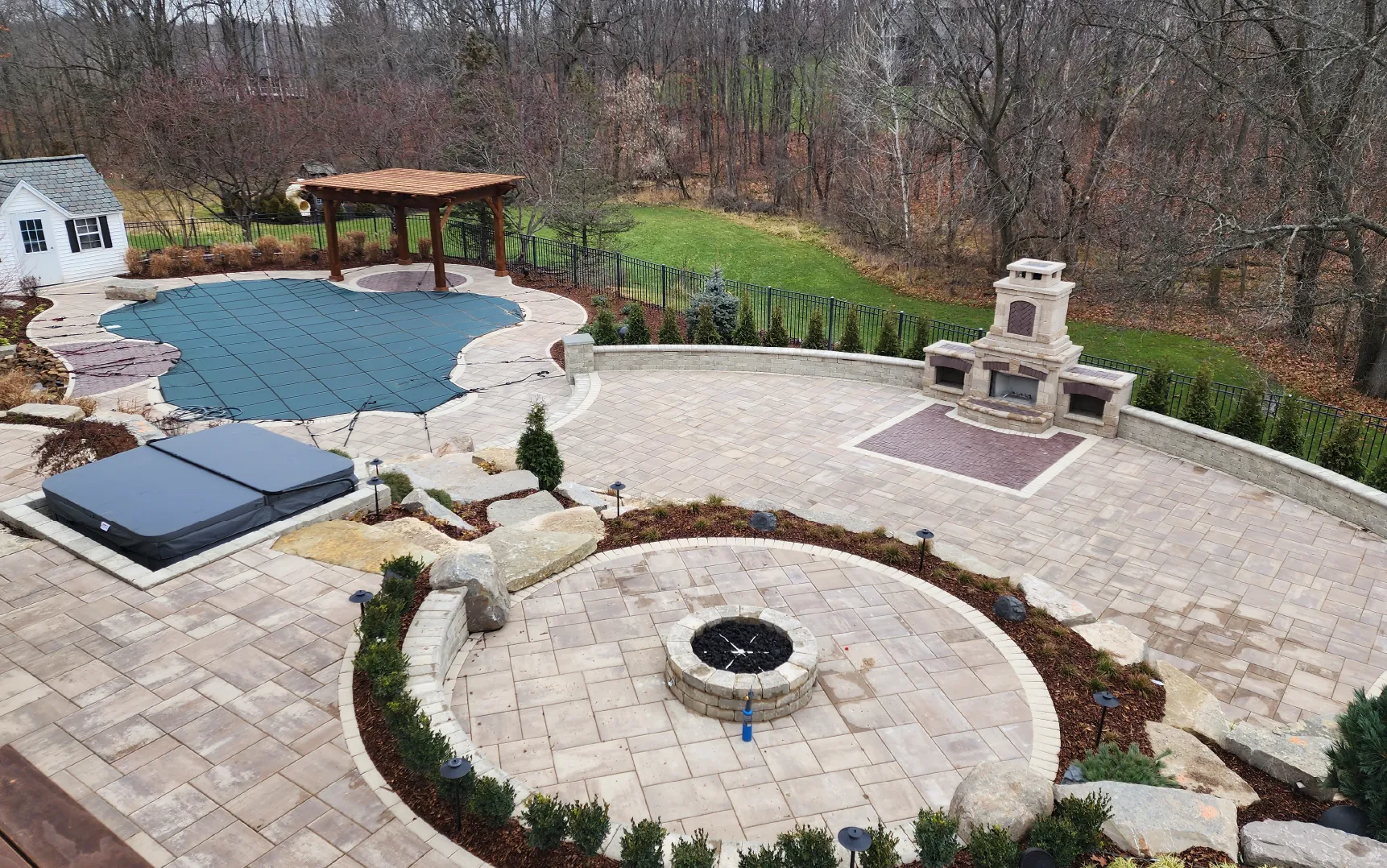 Backyard patio with covered swimming pool, circular fire pit, hot tub, pergola, and stone outdoor fireplace surrounded by trees.